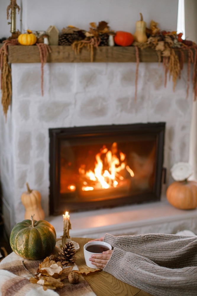 Décoration d'automne cocooning au coin de la cheminée Mains tenant un mug chaud devant une cheminée allumée, décorée de citrouilles et feuilles d'automne.