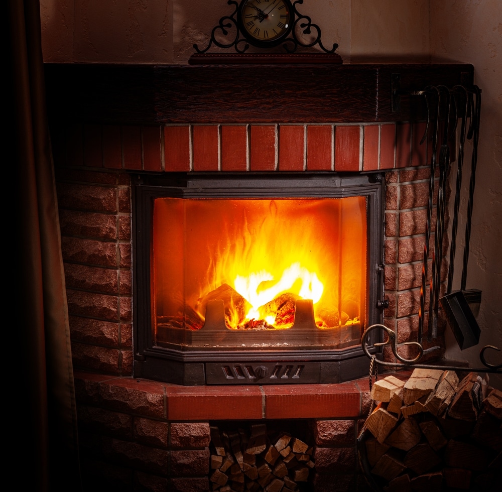 Cheminée en Briques avec Feu de Bois et Ambiance Cosy Cheminée en briques avec un feu vif derrière une vitre, bois de chauffage empilé et horloge décorative ancienne.