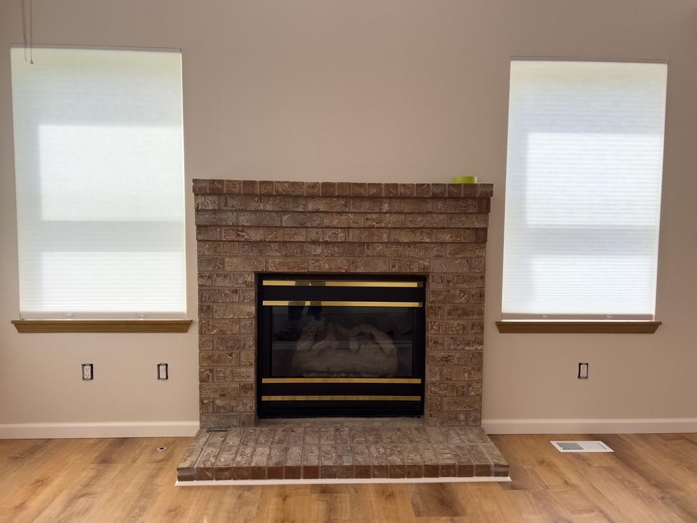 Aménagement Salon : Cheminée en Briques et Grandes Fenêtres Cheminée en briques avec encadrement noir et doré entourée de fenêtres à stores blancs sur sol en bois.