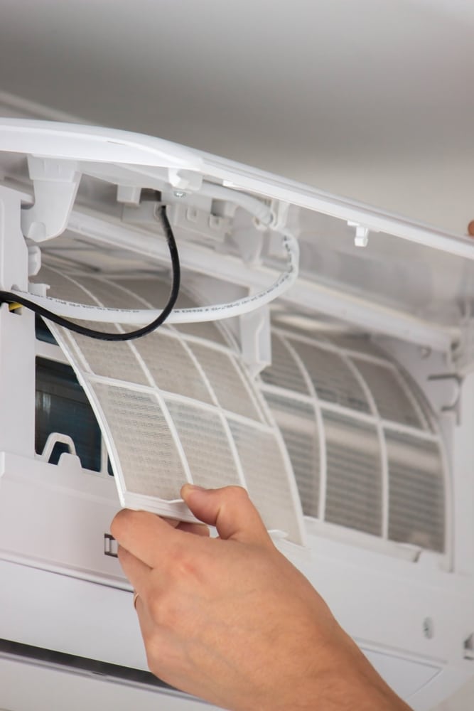 Entretien climatisation : comment nettoyer le filtre à air ? Main retirant un filtre à air poussiéreux d'un climatiseur mural blanc pour un entretien régulier à la maison.