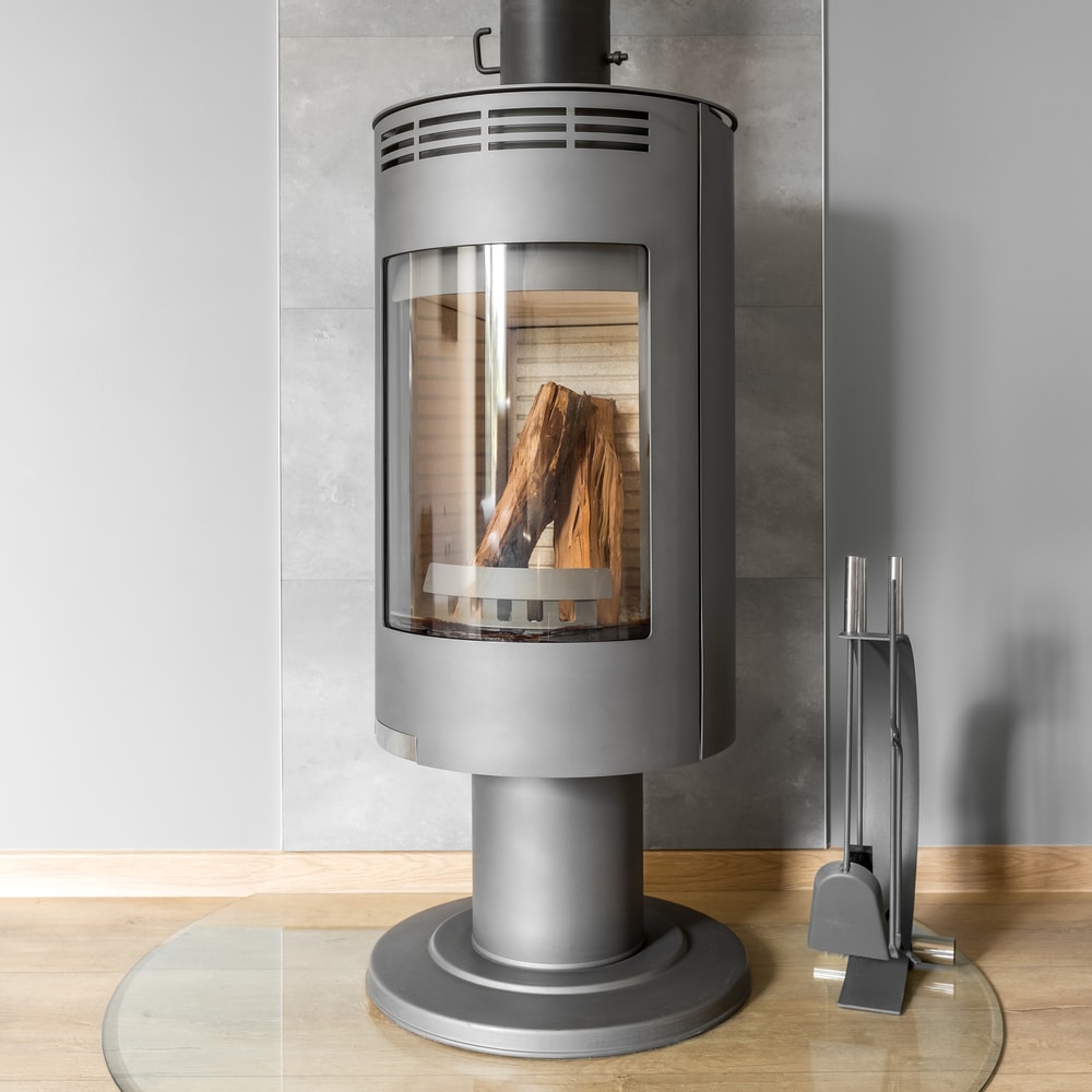 Poêle à Bois Gris Moderne : Chauffage Design et Performant Poêle à bois moderne gris avec bûches derrière sa vitre, posé sur une plaque de protection en verre.
