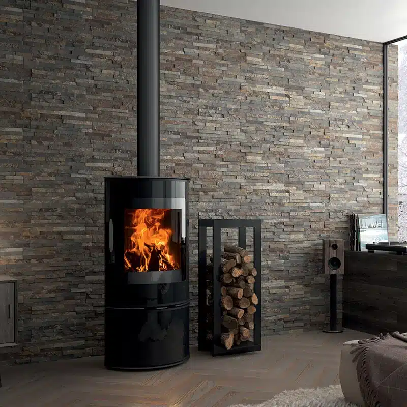 Poêle à bois noir moderne et mur en pierre de parement Poêle à bois cylindrique noir avec feu allumé devant un mur en pierre naturelle et un range-bûches moderne.