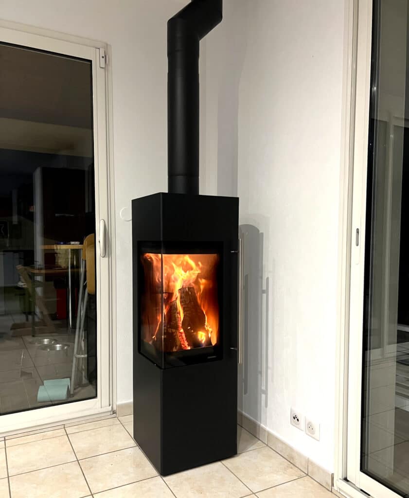 Poêle à bois noir moderne avec vitre d'angle et flammes vives, installé sur un sol en carrelage clair.