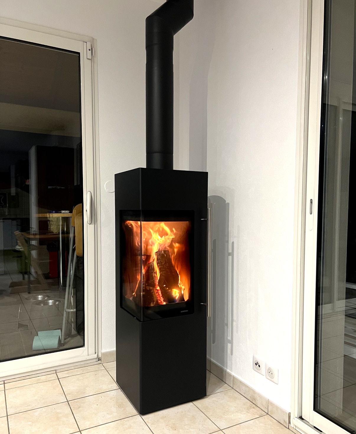 Poêle à bois moderne noir avec vitre d’angle design Poêle à bois noir moderne avec vitre d'angle et flammes vives, installé sur un sol en carrelage clair.